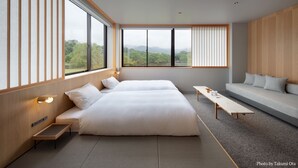 Een moderne hotelkamer met een groot bed, een slaapbank, een tafeltje en grote ramen met uitzicht op groen.