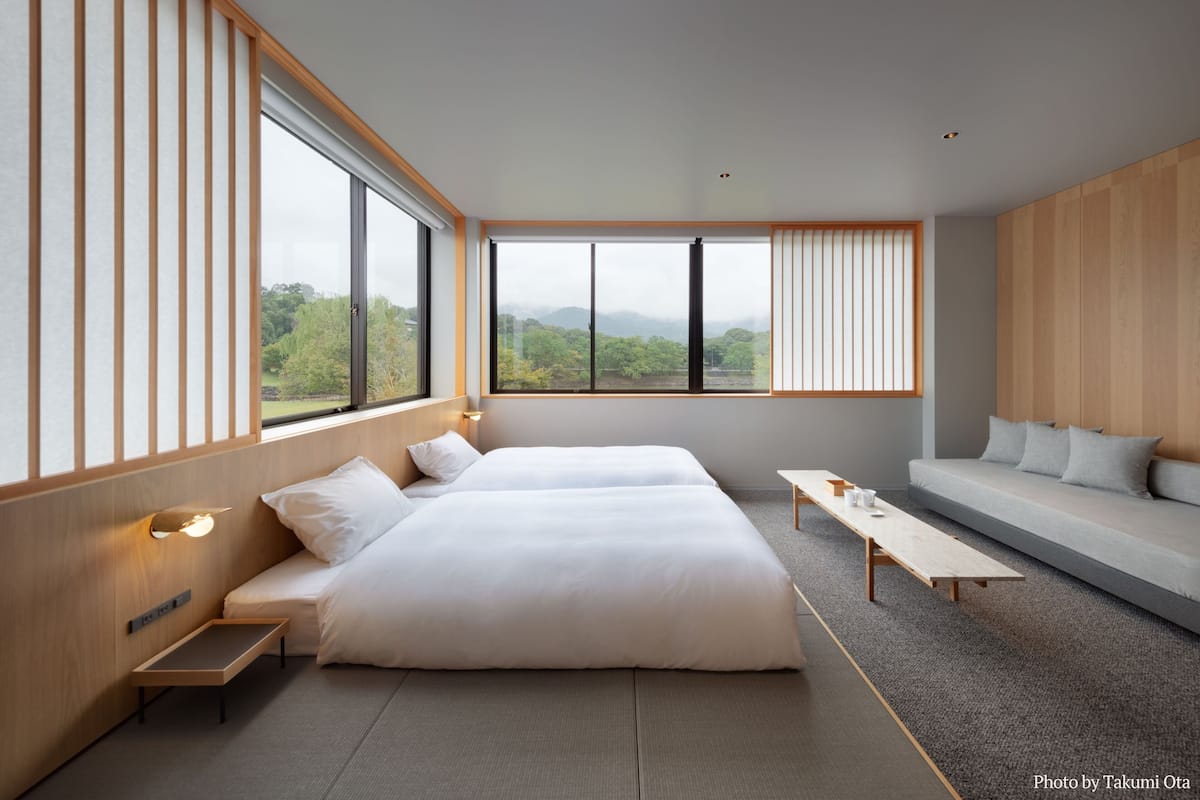 Een moderne hotelkamer met een groot bed, een slaapbank, een tafeltje en grote ramen met uitzicht op groen.