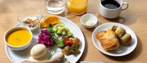 A breakfast plate with soup, sausage, vegetables, and pastries, accompanied by a glass of orange juice and a cup of coffee.