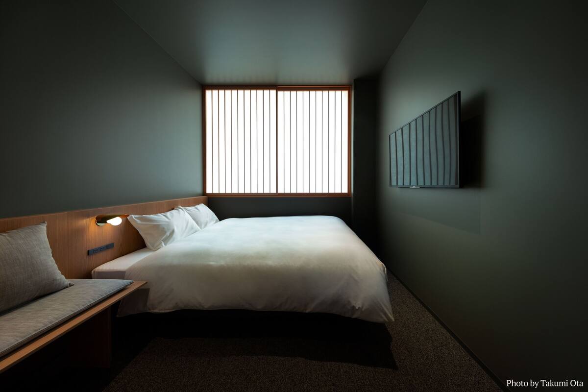 Hotelkamer met een bed, nachtkastje, lamp, raam met zonwering en een kunstwerk aan de muur.