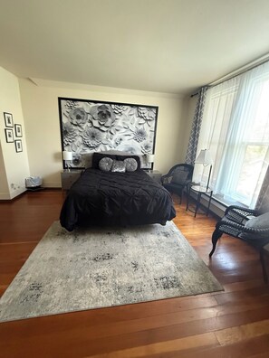 A bedroom with a large bed, a nightstand, a chair, a window with curtains, and a wall with a floral pattern.