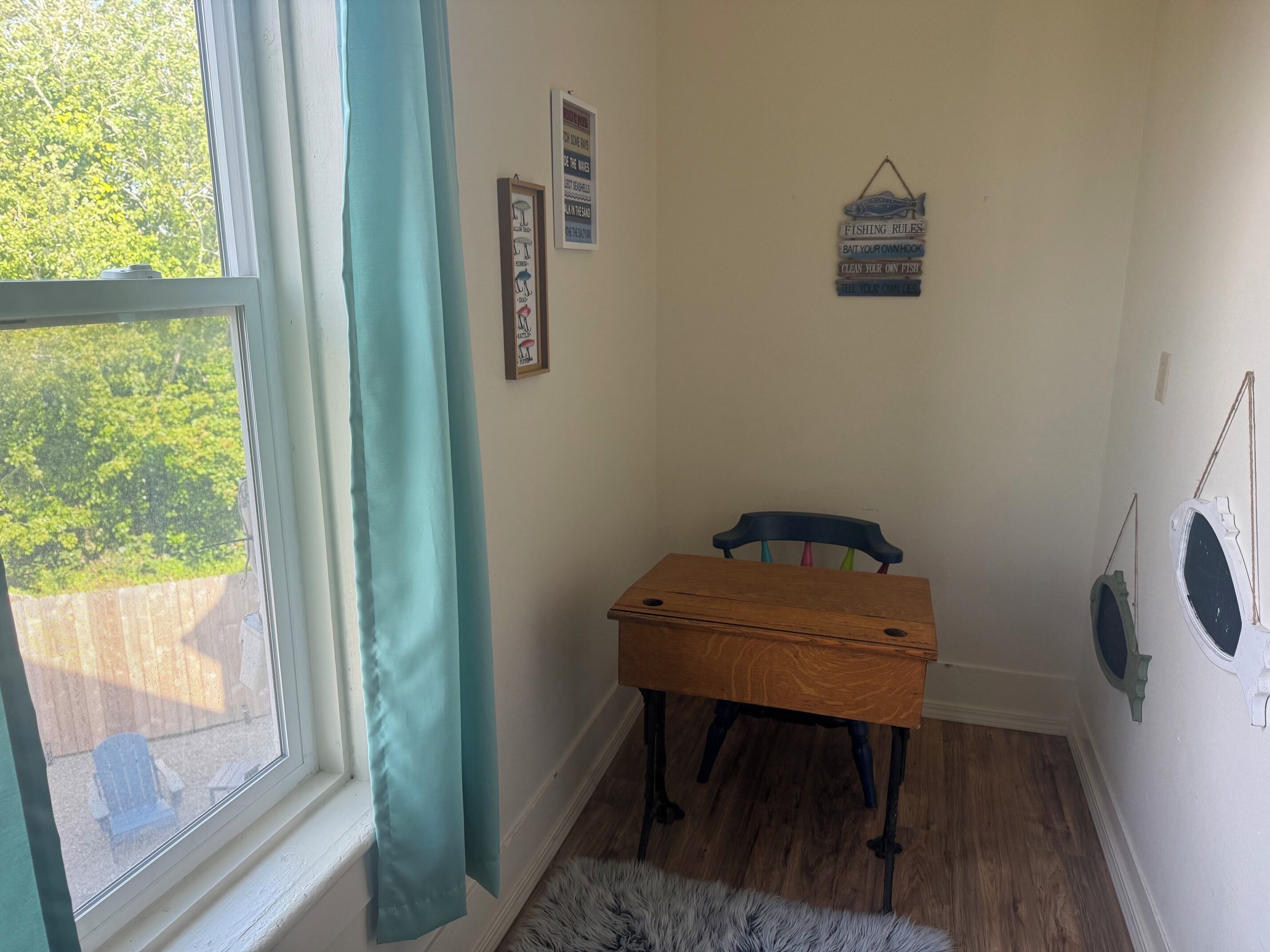 A room with a wooden desk, a chair, a window with a curtain, and a wall with framed items.