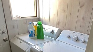 Uma lavanderia compacta com máquina de lavar e secadora frontais, uma janela e produtos de limpeza no balcão.