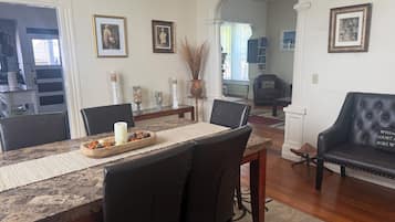A dining area with a long table, chairs, and a buffet tray. There are framed pictures on the walls, a fireplace, and a seating area with a couch.