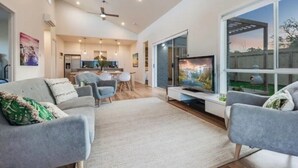 Ein modernes Wohnzimmer mit großem Fernseher, grauen Sofas, einem Essbereich und einer Küche im Hintergrund.