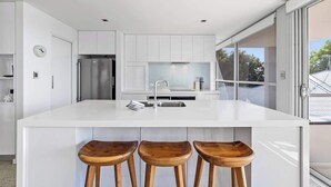 Una cocina moderna con armarios blancos, un refrigerador de acero inoxidable, una isla grande con taburetes y vistas a través de la puerta de vidrio.