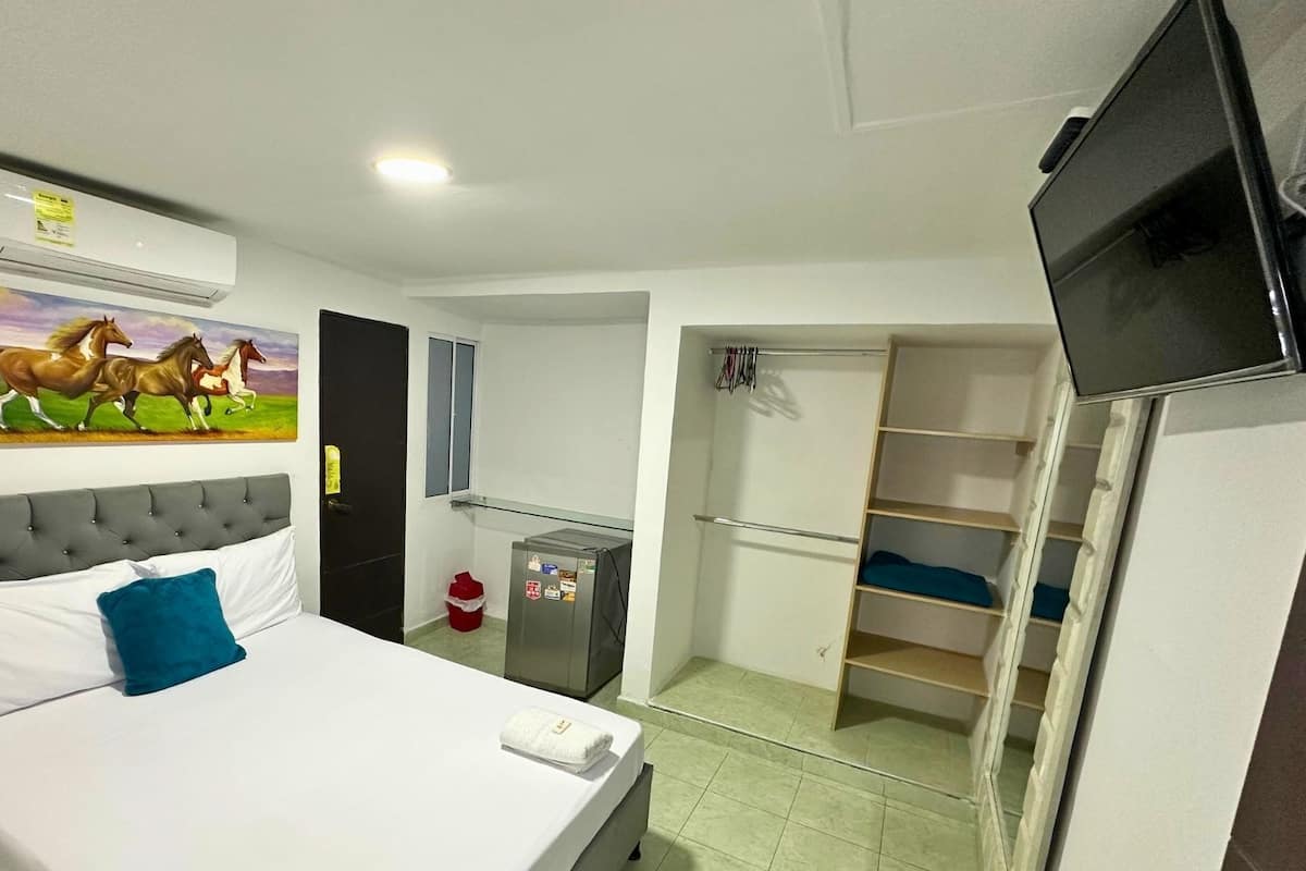 A hotel room with a bed, a television, a painting of horses, a wardrobe, and a small refrigerator.