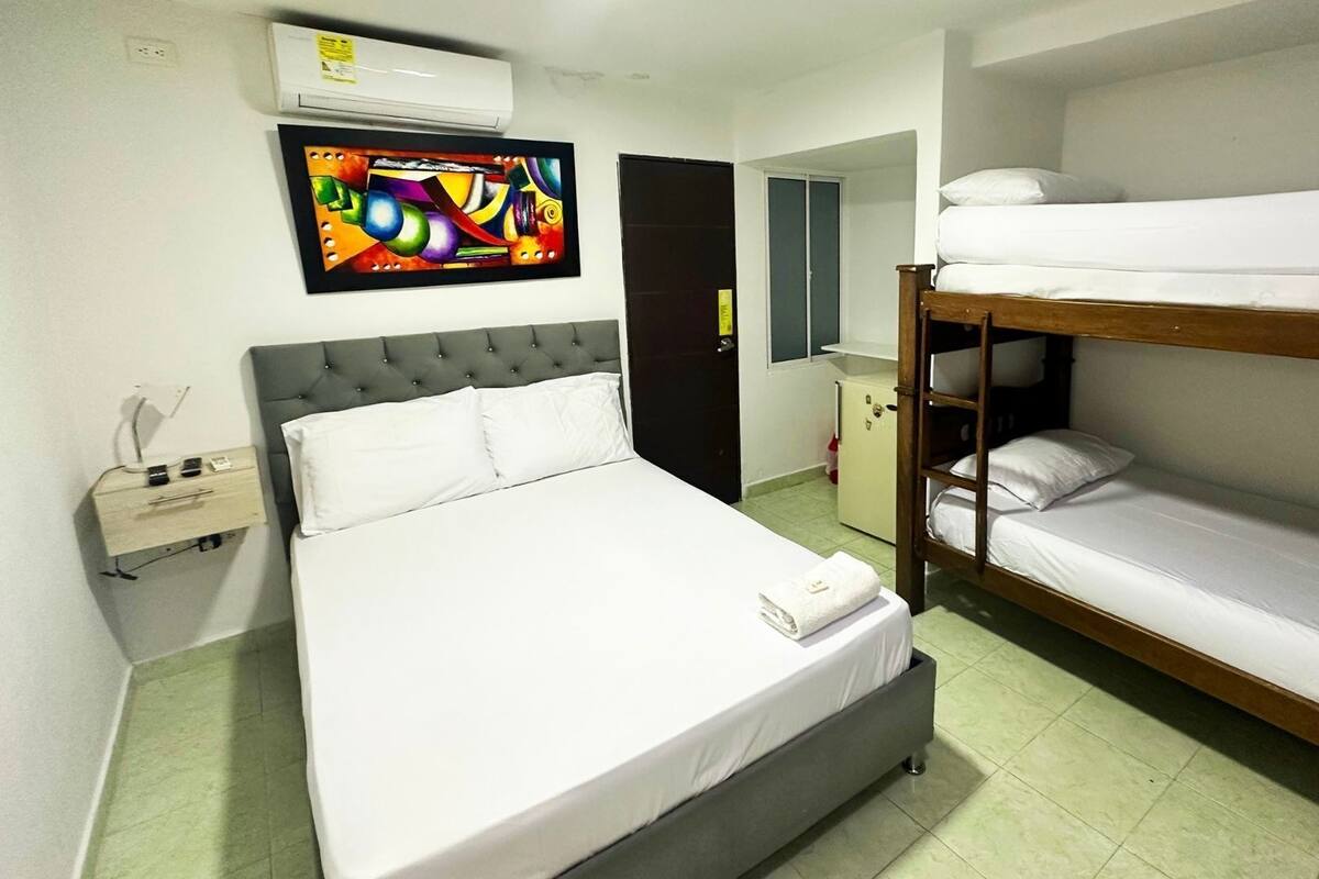 A hotel room with a single bed, a bunk bed, a bedside table with a lamp, a wall-mounted air conditioner, and a colorful painting.