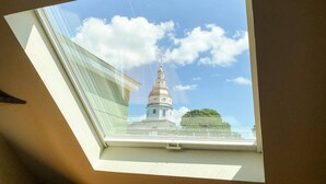 Une lucarne offrant une vue sur un bâtiment blanc doté d’un dôme et d’une flèche, sous un ciel bleu dégagé.
