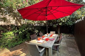 Una mesa de comedor con sillas rojas y un paraguas rojo, rodeada de follaje y un muro de ladrillo.