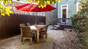 Une terrasse avec une table et des chaises en bois, un parasol rouge et un chemin en briques.