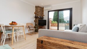A living room with a stone wall, a television, a sofa, a wooden table with fruit, and a balcony with a view. - Adelfa 7 by Interhome (L'Escala)