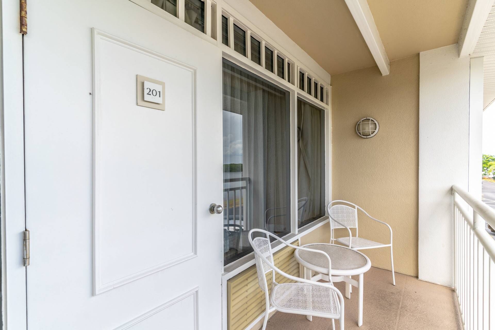 Un balcone con due sedie in metallo bianco e un tavolo, una porta bianca con una targa numerica e una finestra con tende.