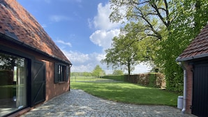 A courtyard with cobblestone flooring, a brick building with a tiled roof, and a grassy area with trees. - Holiday home surrounded by woods and meadows (Grote Brogel)