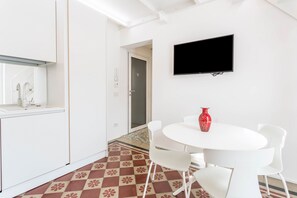 Une cuisine moderne avec des meubles blancs, un sol carrelé, un coin repas avec une table et des chaises blanches, et un téléviseur à écran plat fixé au mur.