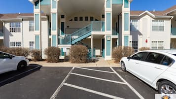 Un immeuble à plusieurs étages doté d’un escalier central, d’un parking et de deux voitures blanches garées devant.