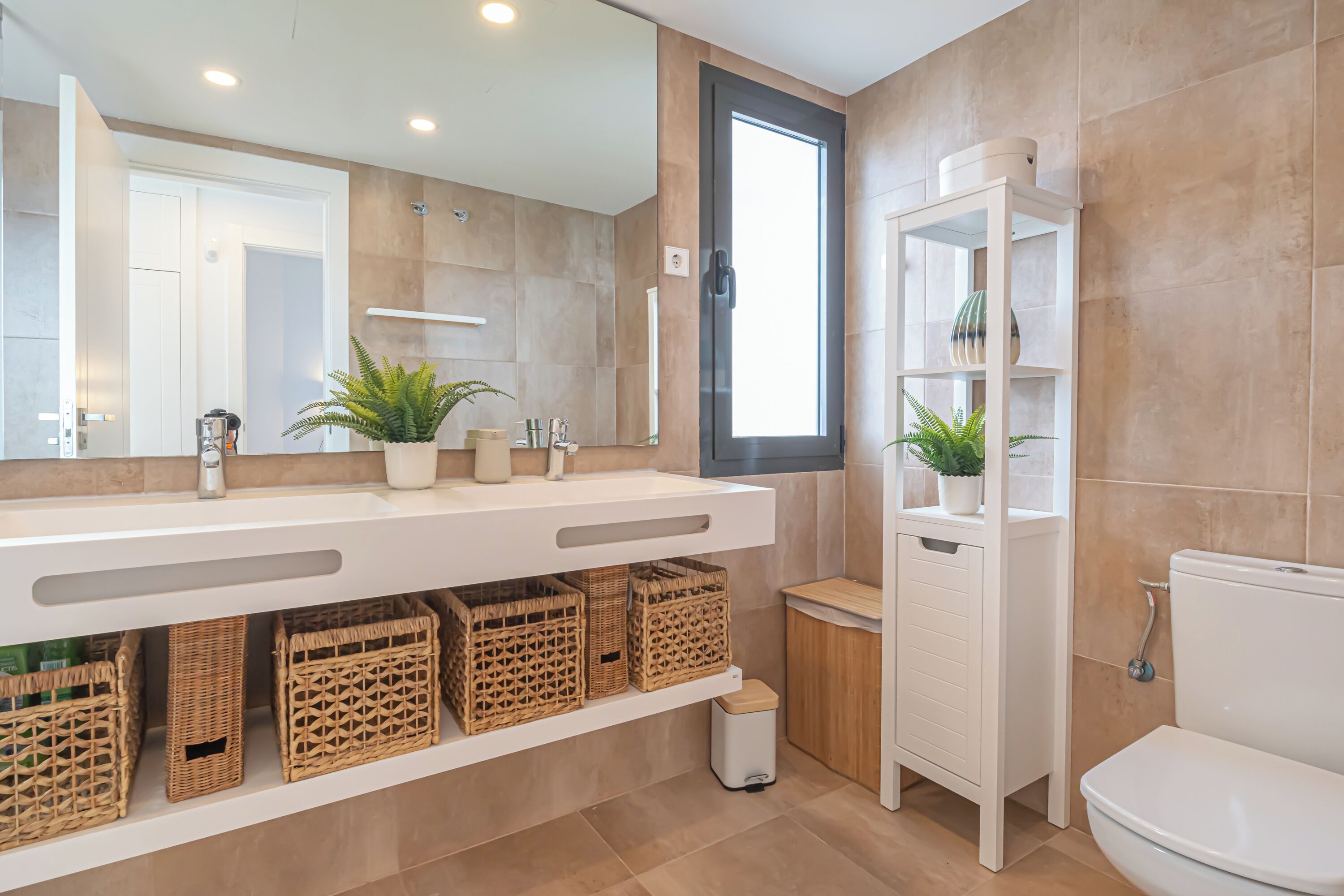 Un baño moderno con un espejo grande, tocador blanco, cestas de mimbre y un inodoro.