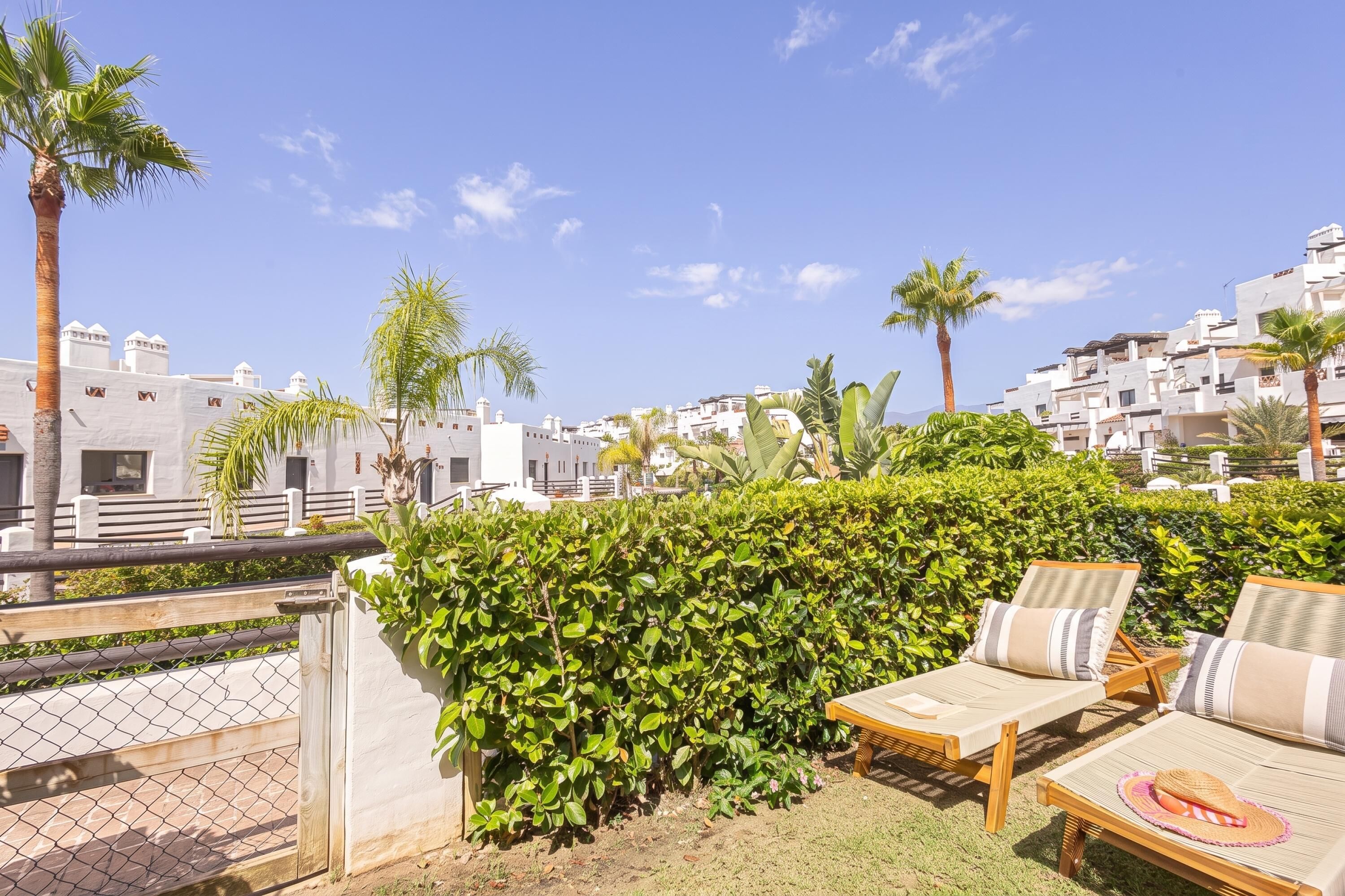 A rooftop terrace with lounge chairs, a wooden table, and a view of white buildings and palm trees.