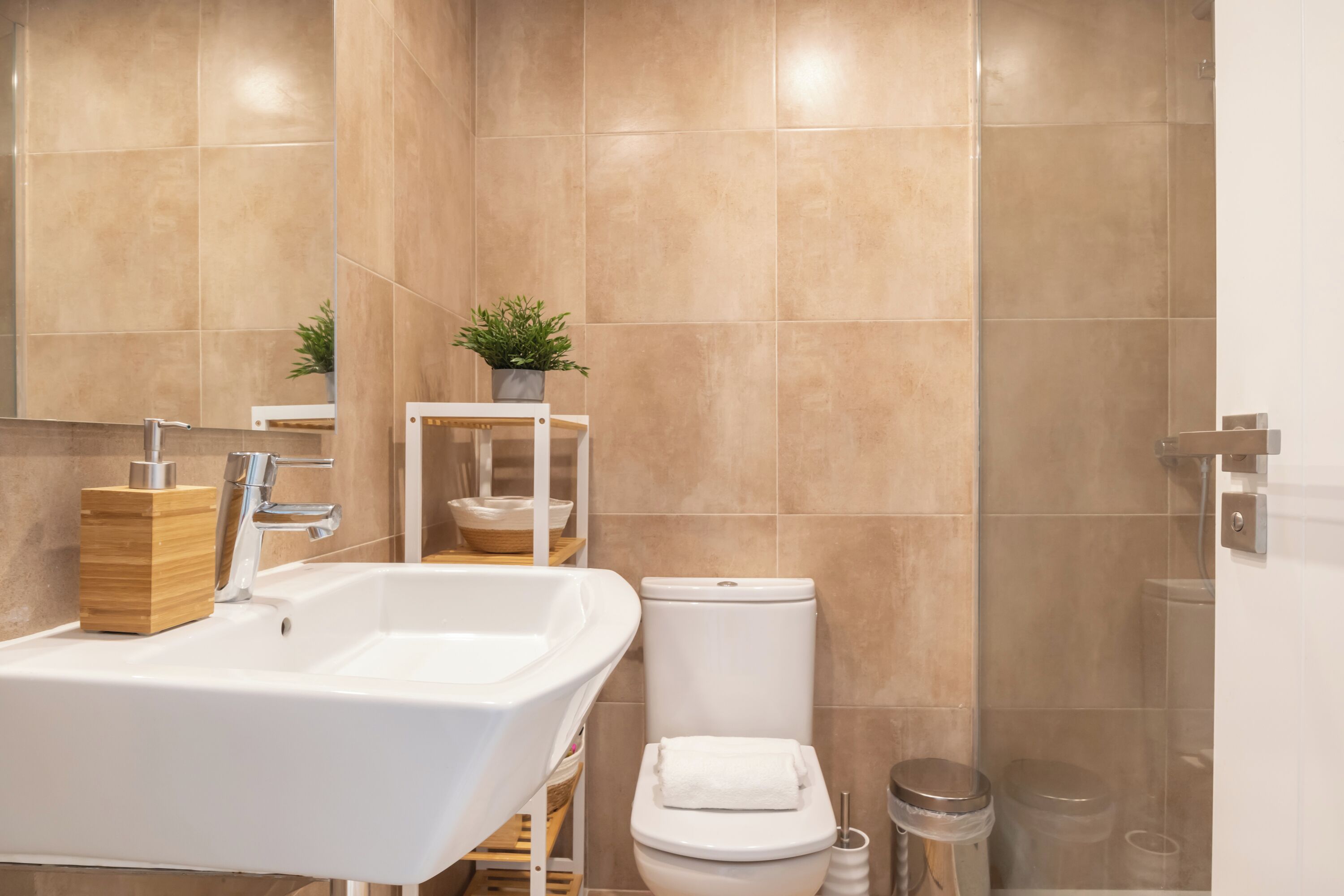 A modern bathroom with a white sink, a wooden cabinet, a toilet, and a small plant.