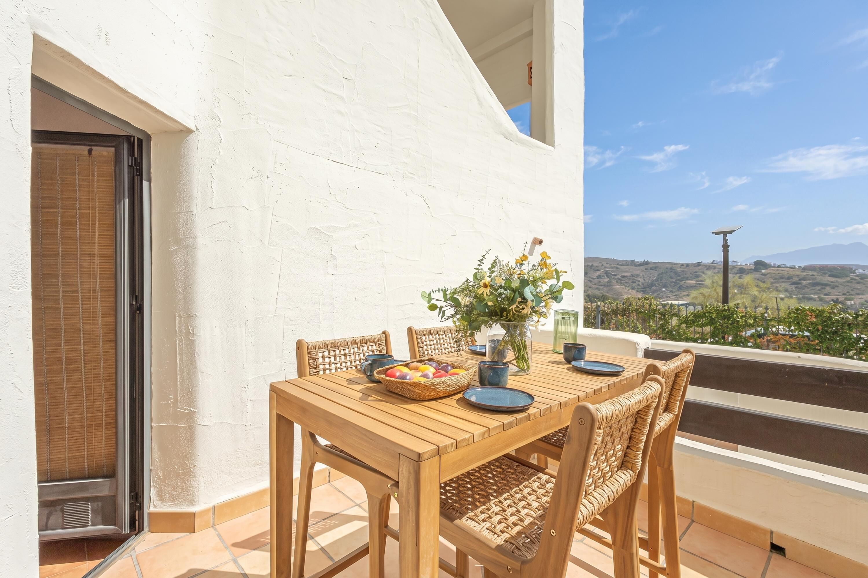A wooden table with chairs, a vase with flowers, and a bowl of fruit on a balcony with a white wall and a view of the landscape.