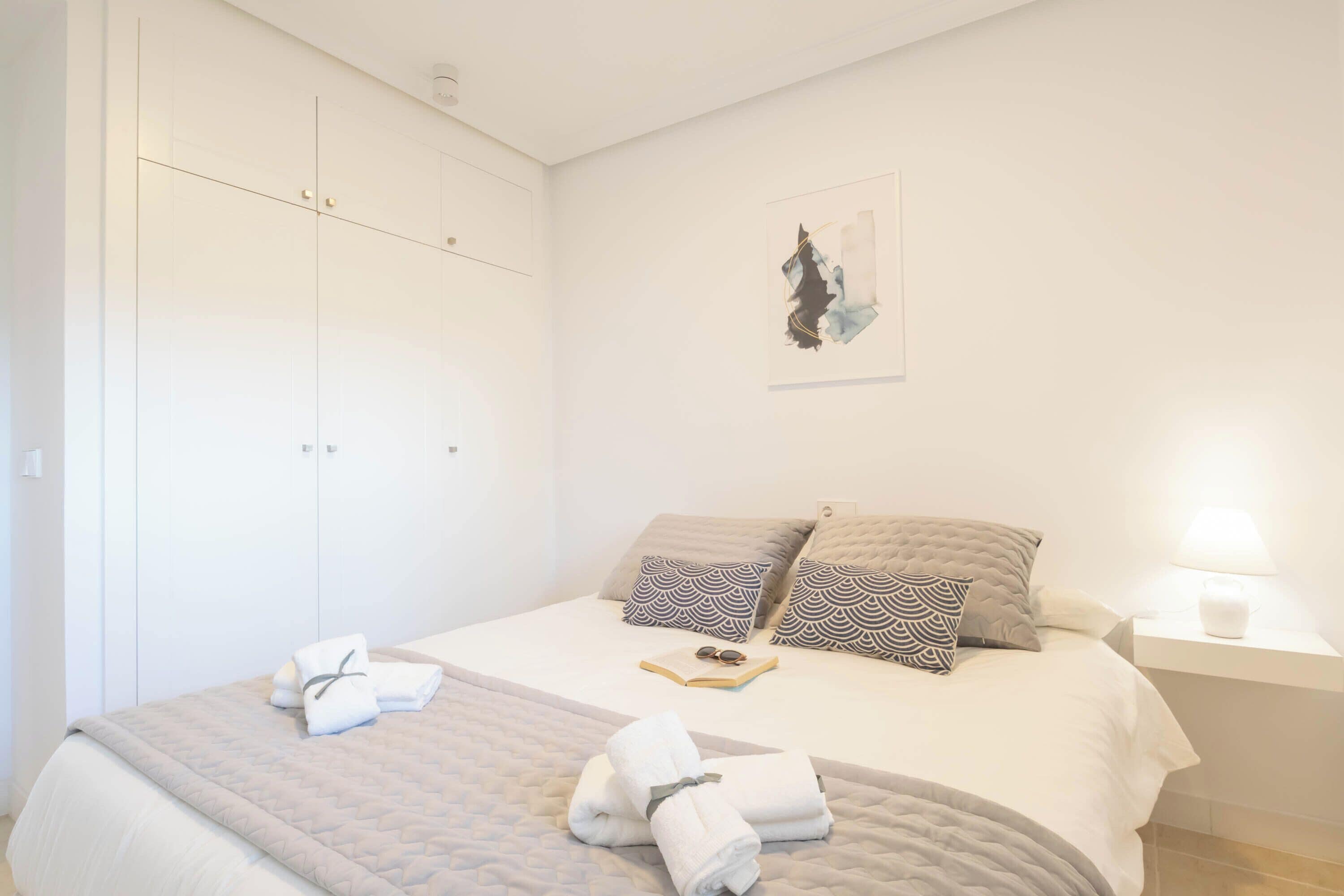 A neatly made bed with a quilt, pillows, and folded towels. There is a bedside table with a lamp and a framed artwork on the wall.