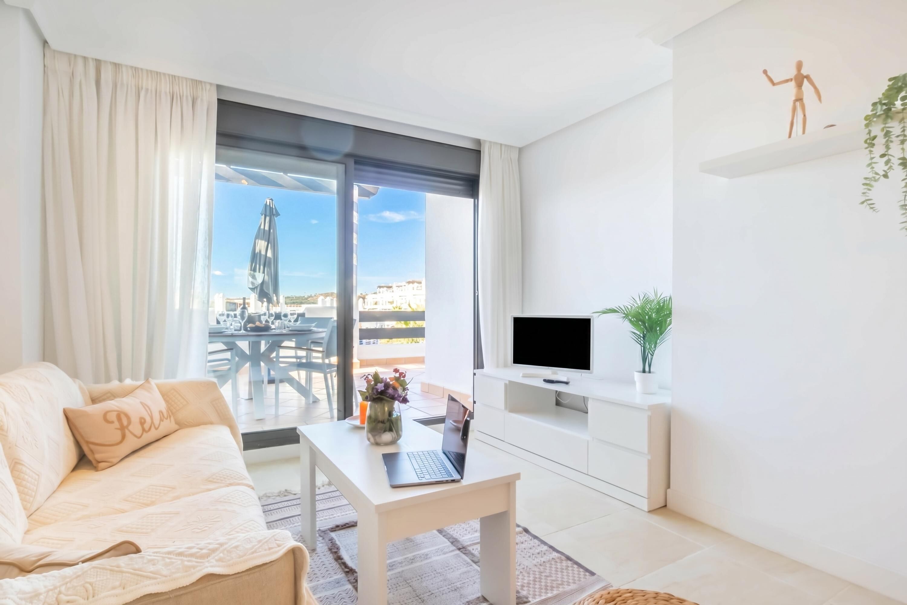 Een moderne woonkamer met een bank, een tafel met een laptop, een tv op een standaard, en een balkon met uitzicht op gebouwen en een buitentafel.