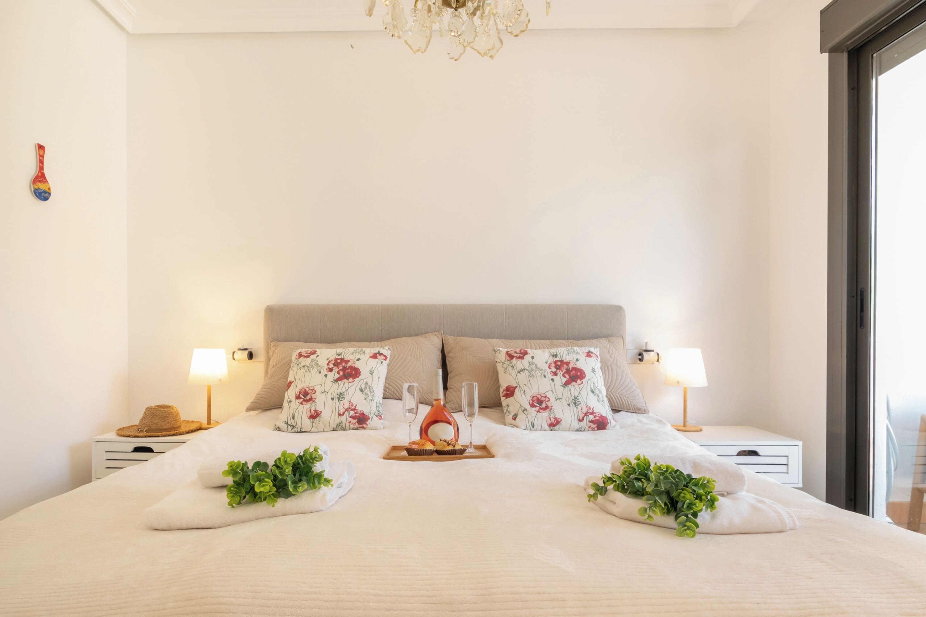 A neatly made bed with floral pillows, bedside lamps, and a tray with a bottle and glasses.