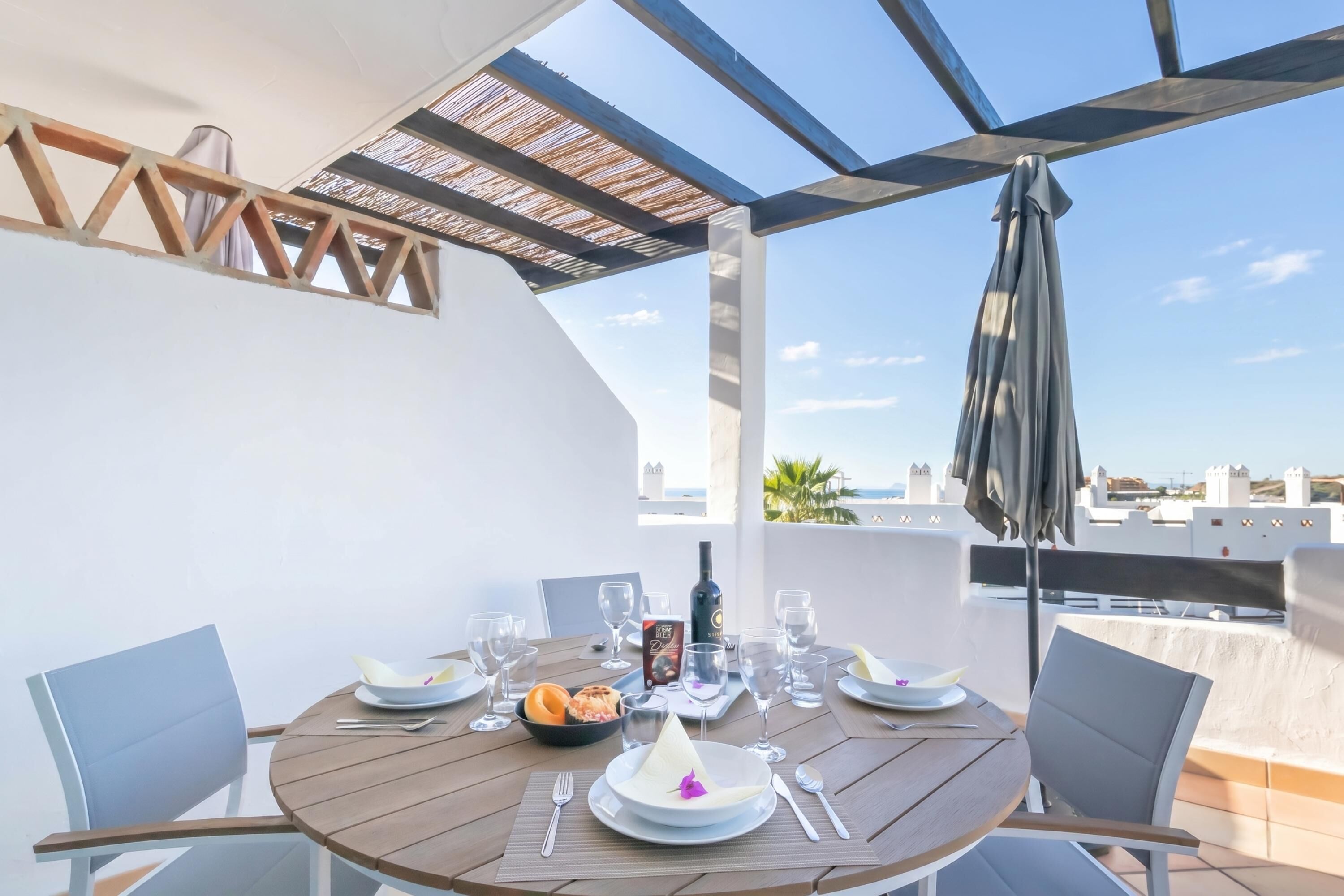 Ein runder Tisch mit Stühlen, einer Flasche, Gläsern und einer Obstschale auf einer Terrasse mit Blick.
