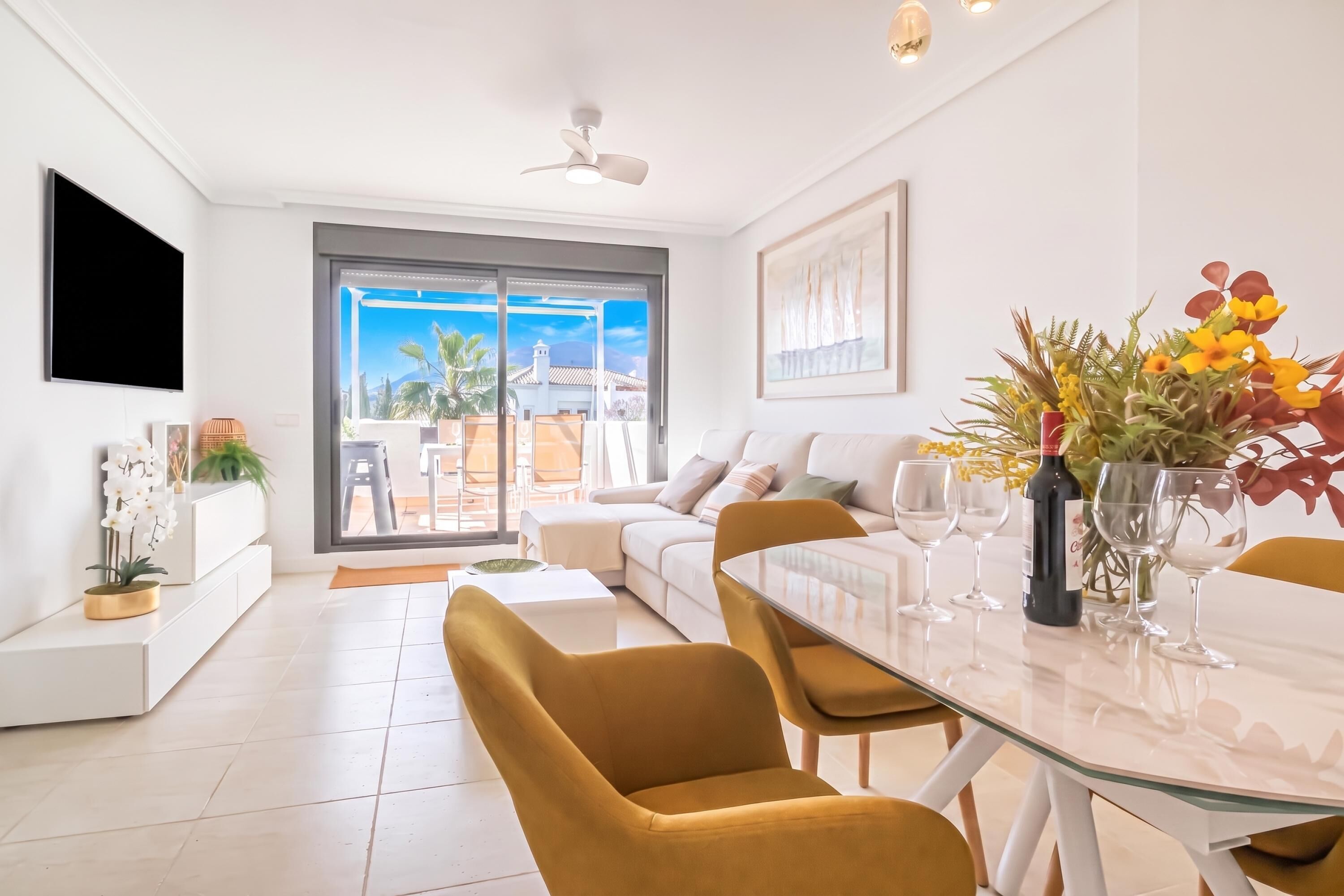 A modern living room with a glass dining table, comfortable seating, a flat-screen TV, and a view of the outdoors.