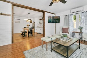 A spacious living room with a wooden floor, a dining area with a wooden table and chairs, a sofa, and a coffee table. There is a painting on the wall, a ceiling fan, and an air conditioning unit.