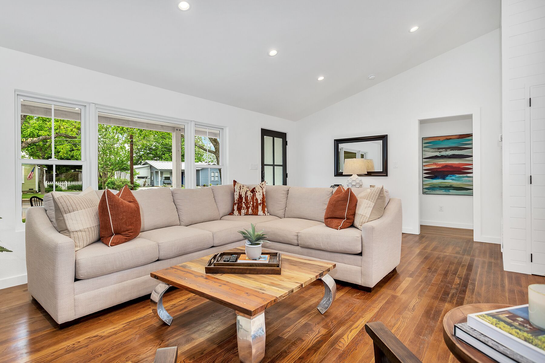 Un salon moderne avec un canapé d’angle, une table basse en bois et une grande fenêtre donnant sur une maison et un espace verdoyant.