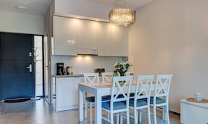 A modern kitchen with white cabinets, a dining table with four chairs, a coffee machine, and a chandelier.