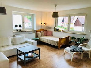 Un salon chaleureux avec un canapé, une petite table, une banquette et un fauteuil. De grandes fenêtres offrent une vue sur la verdure et un bâtiment situé à l’extérieur.