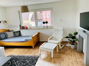 Un salon moderne avec un lit en bois, un fauteuil blanc, une petite table, un téléviseur et une fenêtre donnant sur des immeubles.