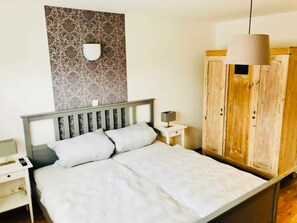 A bed with a headboard, a bedside table with a lamp, a wall-mounted light fixture, and a wooden wardrobe.