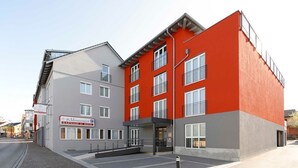 Un hotel moderno con fachada roja y gris, múltiples ventanales y una entrada visible.