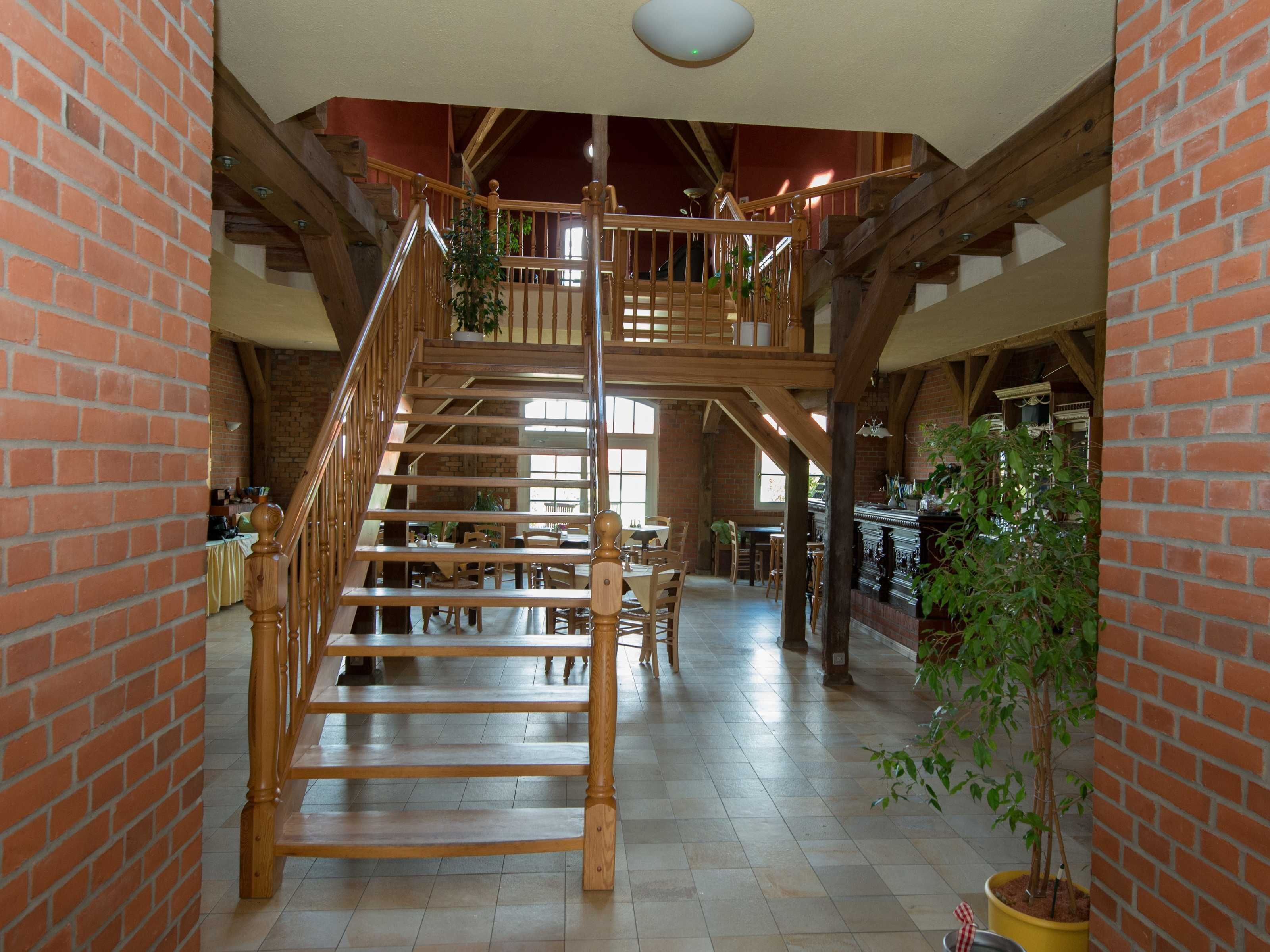 Un interior amplio con una escalera de madera que conduce a un nivel superior, un comedor con mesas y sillas, y una pared de ladrillo en un lateral.