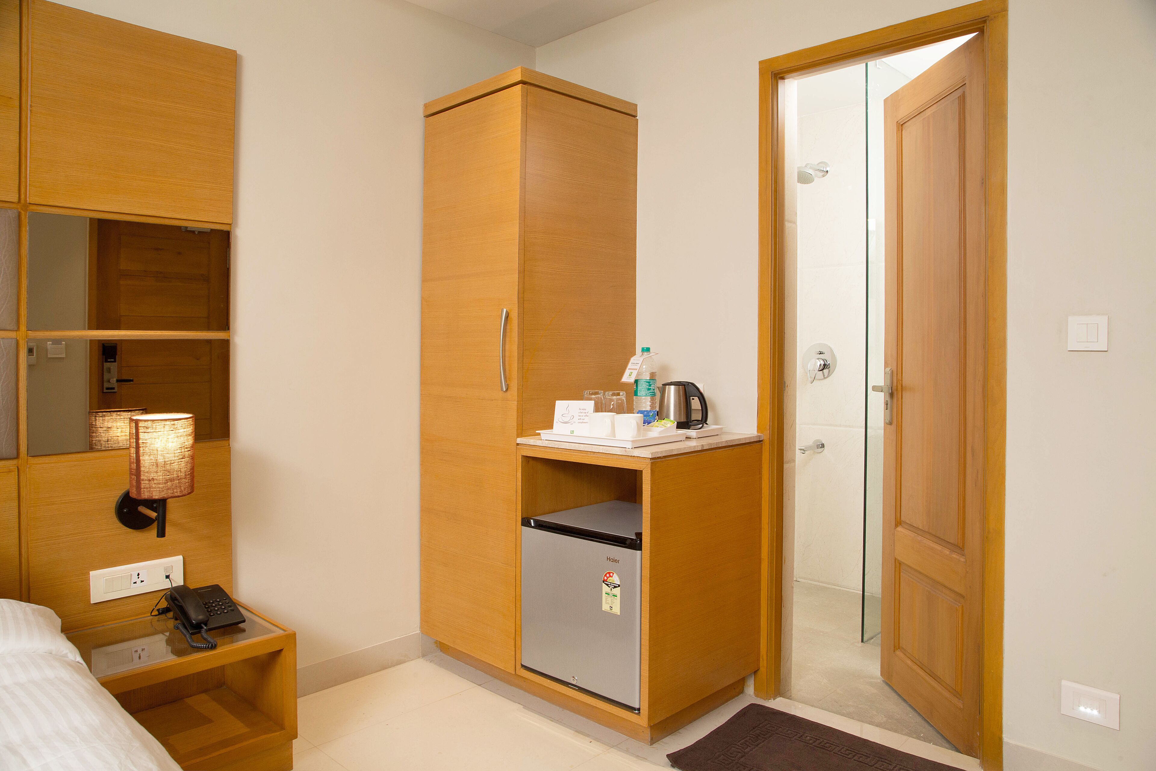 A hotel room with a bed, a wooden wardrobe, a small refrigerator, a nightstand with a phone, and a bathroom door.