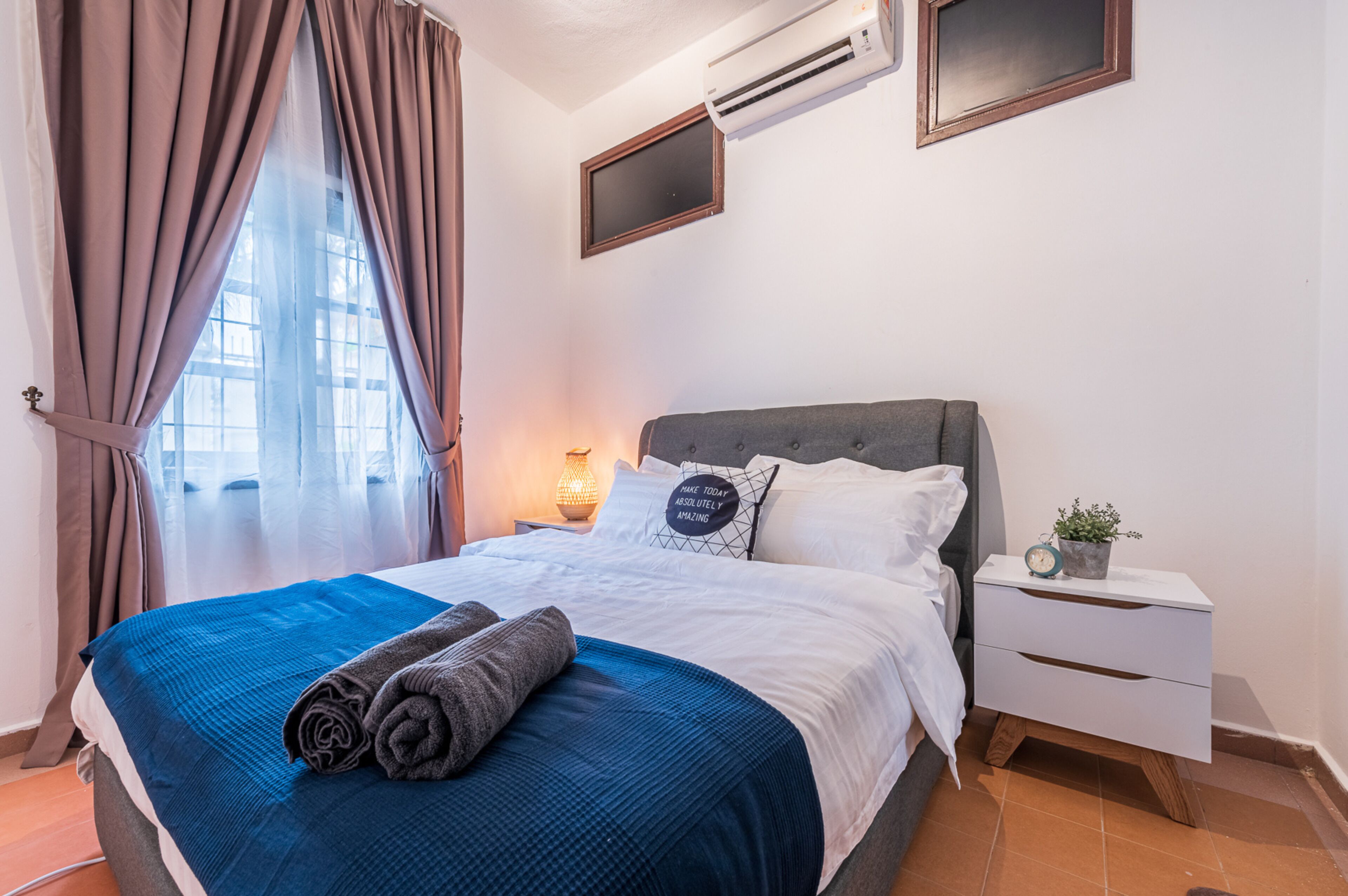 A neatly made bed with a blue blanket and rolled towels, a bedside table with a clock and a plant, and a window with curtains.