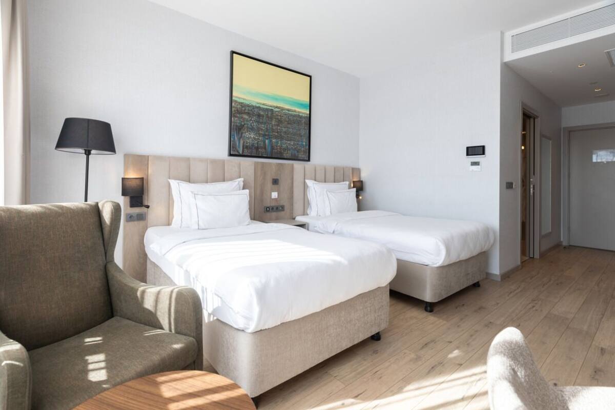 Habitación de hotel con dos camas, un sofá, una lámpara y un cuadro de un paisaje urbano en la pared.
