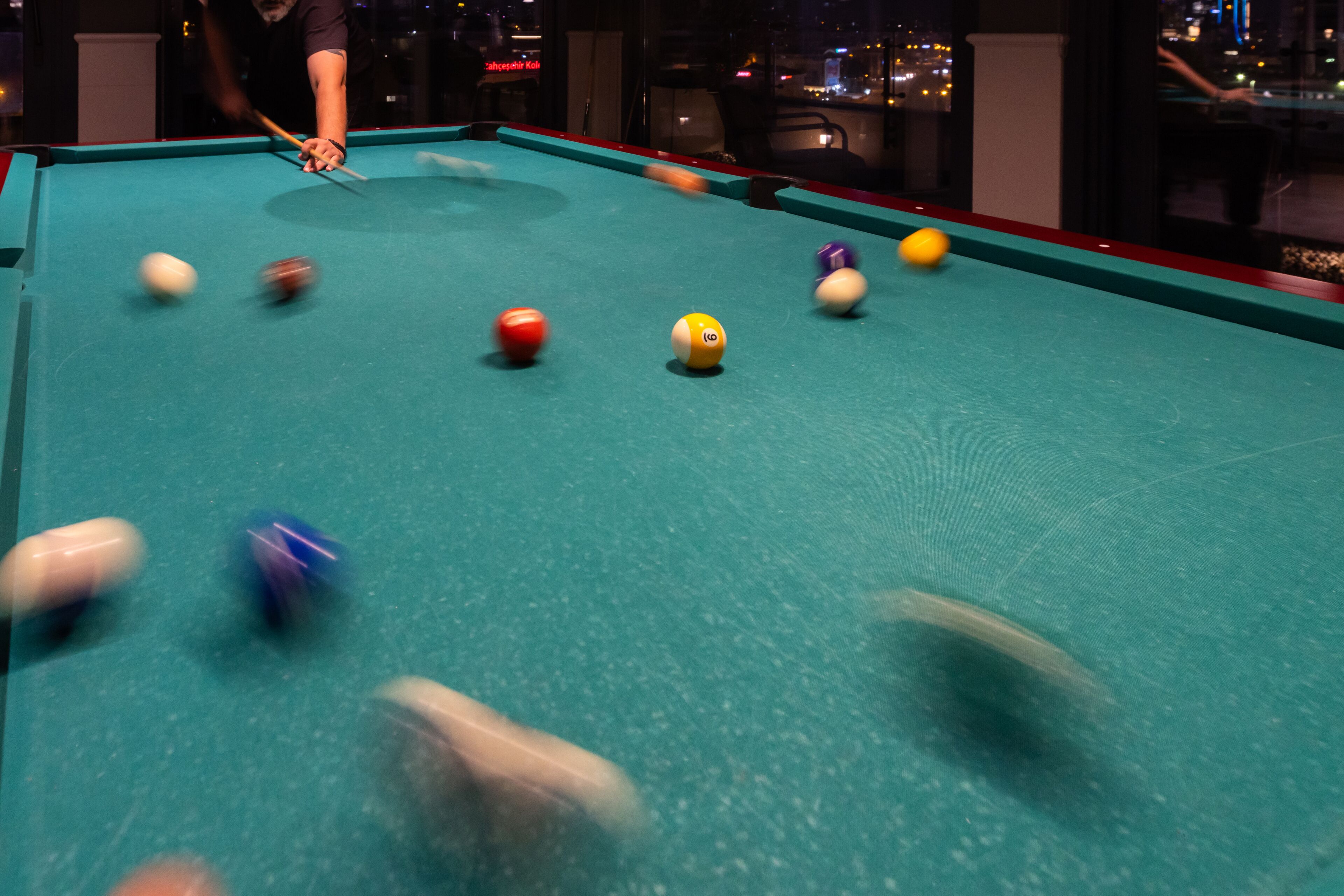 A pool table with balls in motion, a pool cue, and a person playing.