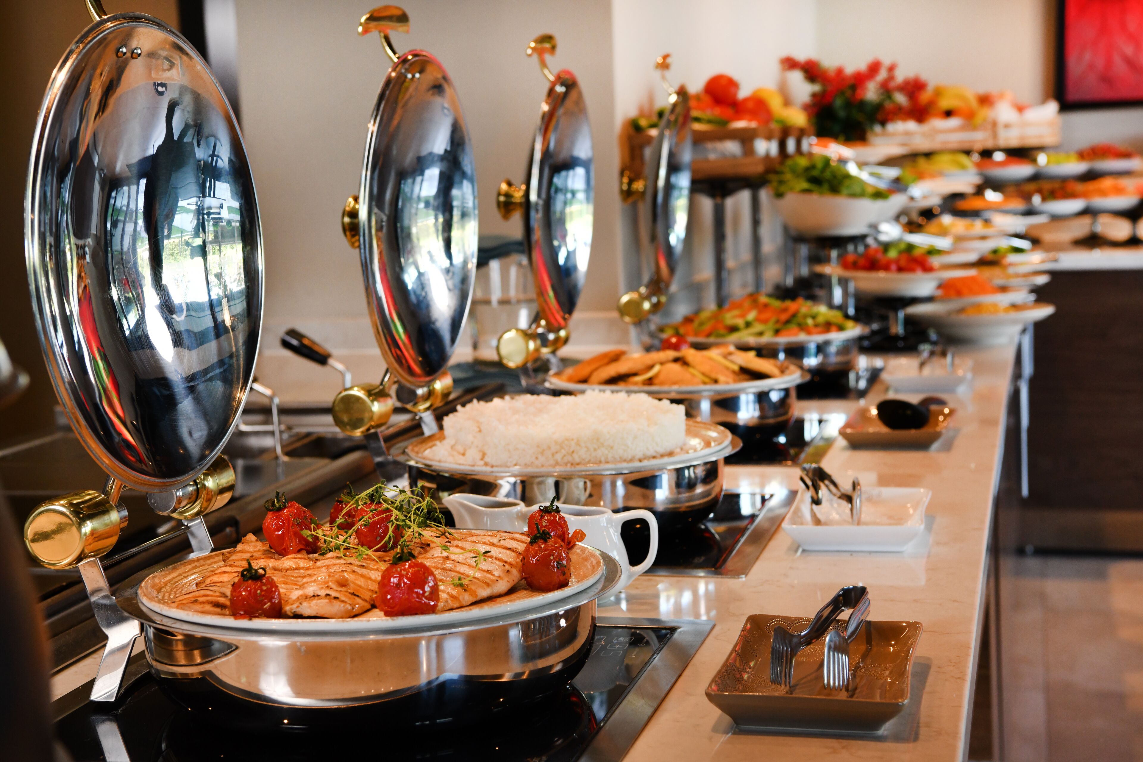 A buffet with various dishes including rice, salads, and grilled chicken, with reflective serving dishes and utensils.