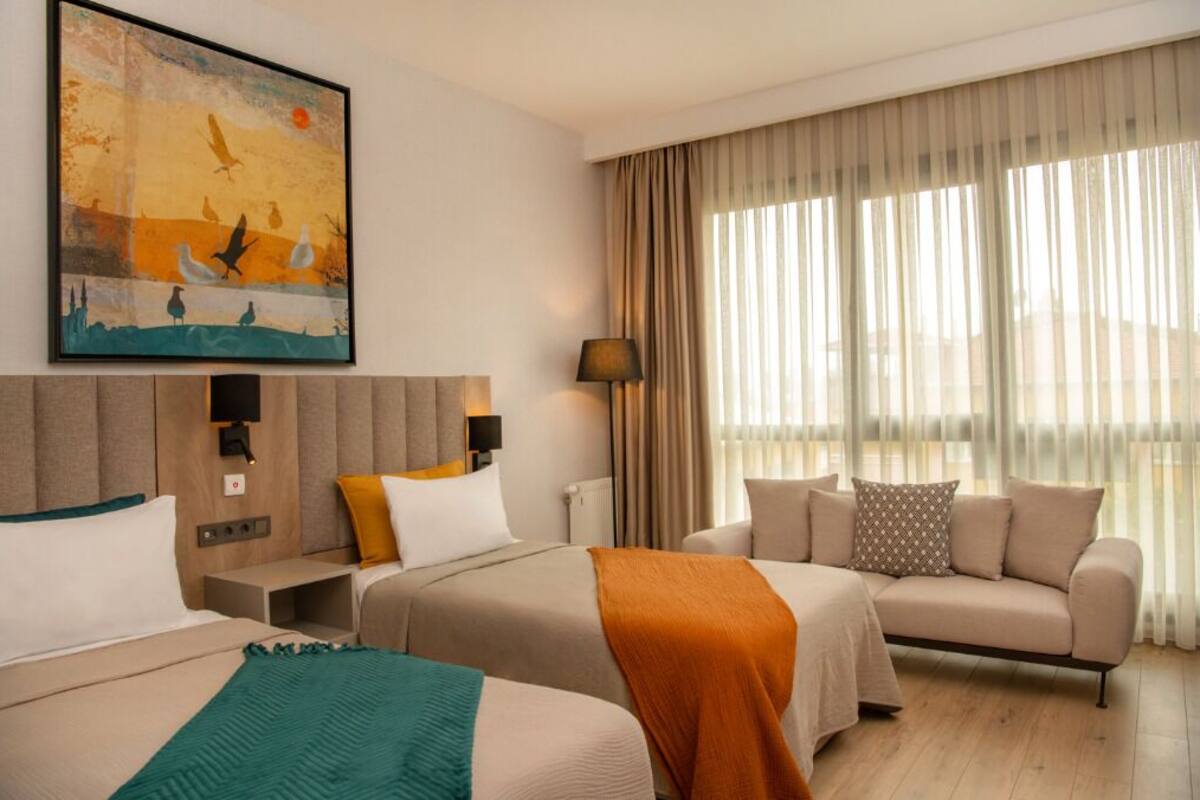 Habitación de hotel con dos camas, un sofá, un cuadro, una lámpara y un ventanal grande con cortinas.