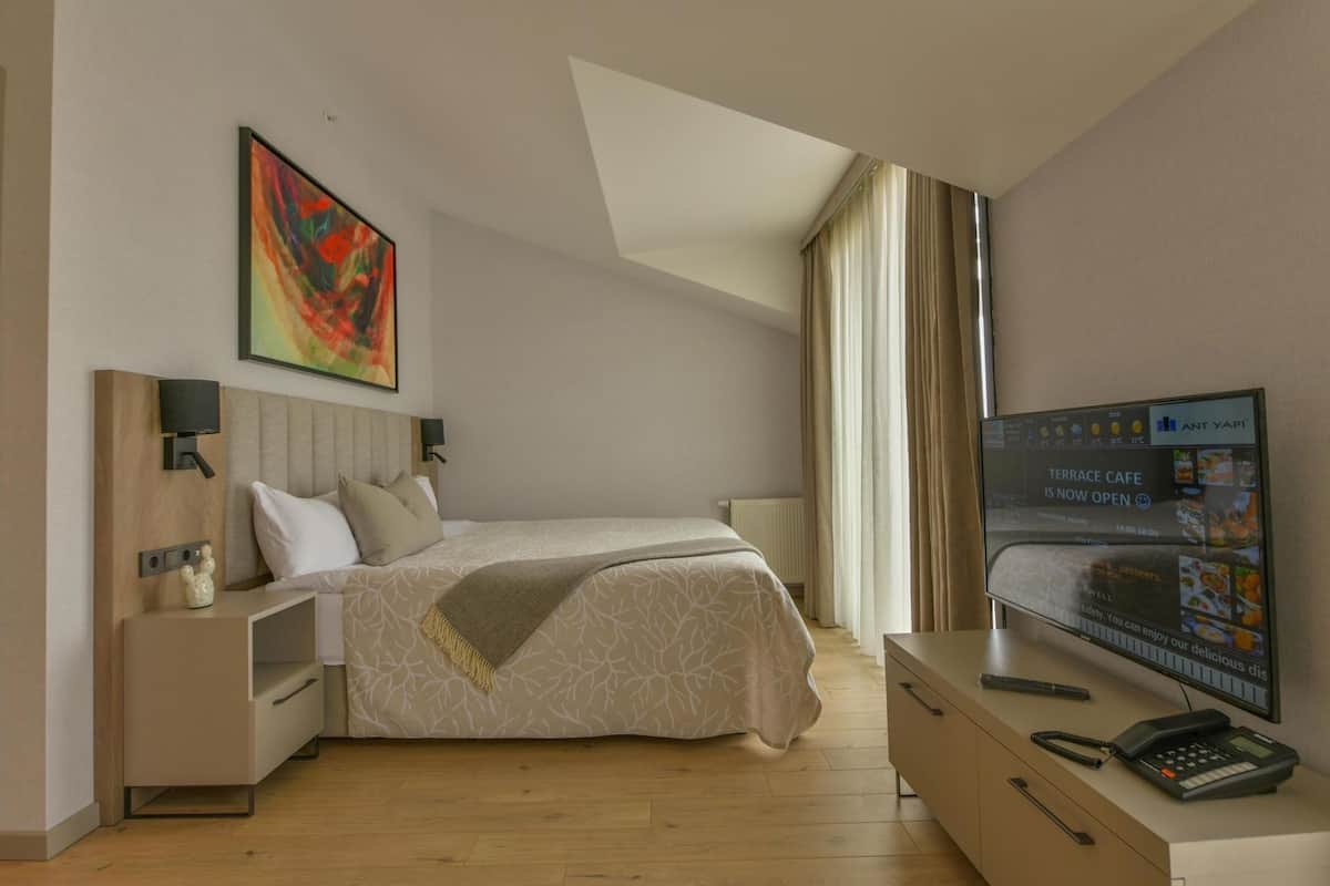 Una habitación de hotel con cama, mesitas de noche, televisión y ventana con cortinas.