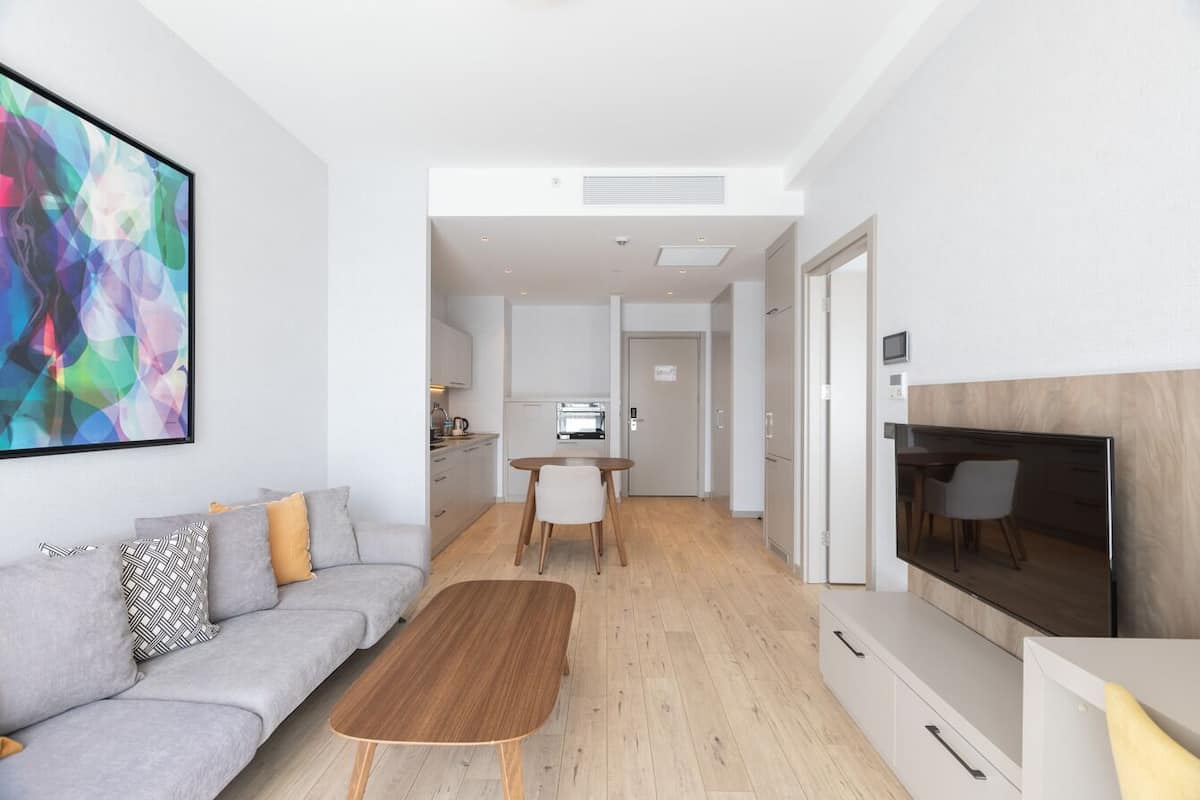 Una sala de estar moderna con un sofá gris, una mesa de centro de madera, un televisor y una cocina al fondo.