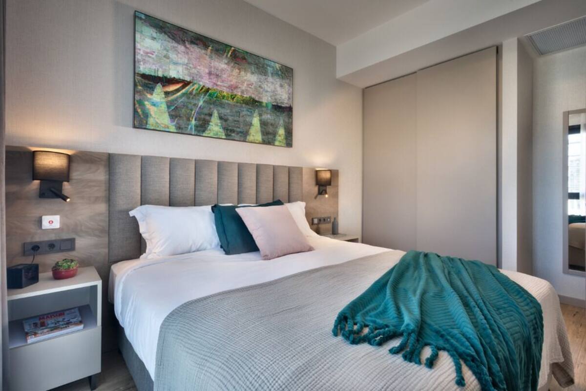 Una habitación de hotel moderna con una cama grande, un cabecero con iluminación integrada, una mesita de noche con lámpara y un cuadro en la pared.