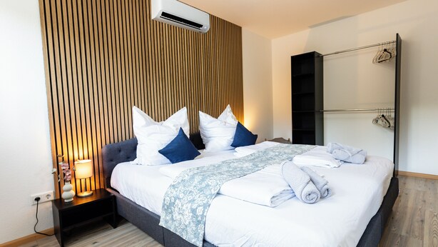 A modern hotel room with a large bed, a bedside table with a lamp, a wardrobe, and an air conditioning unit.