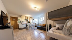 A modern kitchen with wooden flooring, a dining area with chairs, and a sofa with a fan.