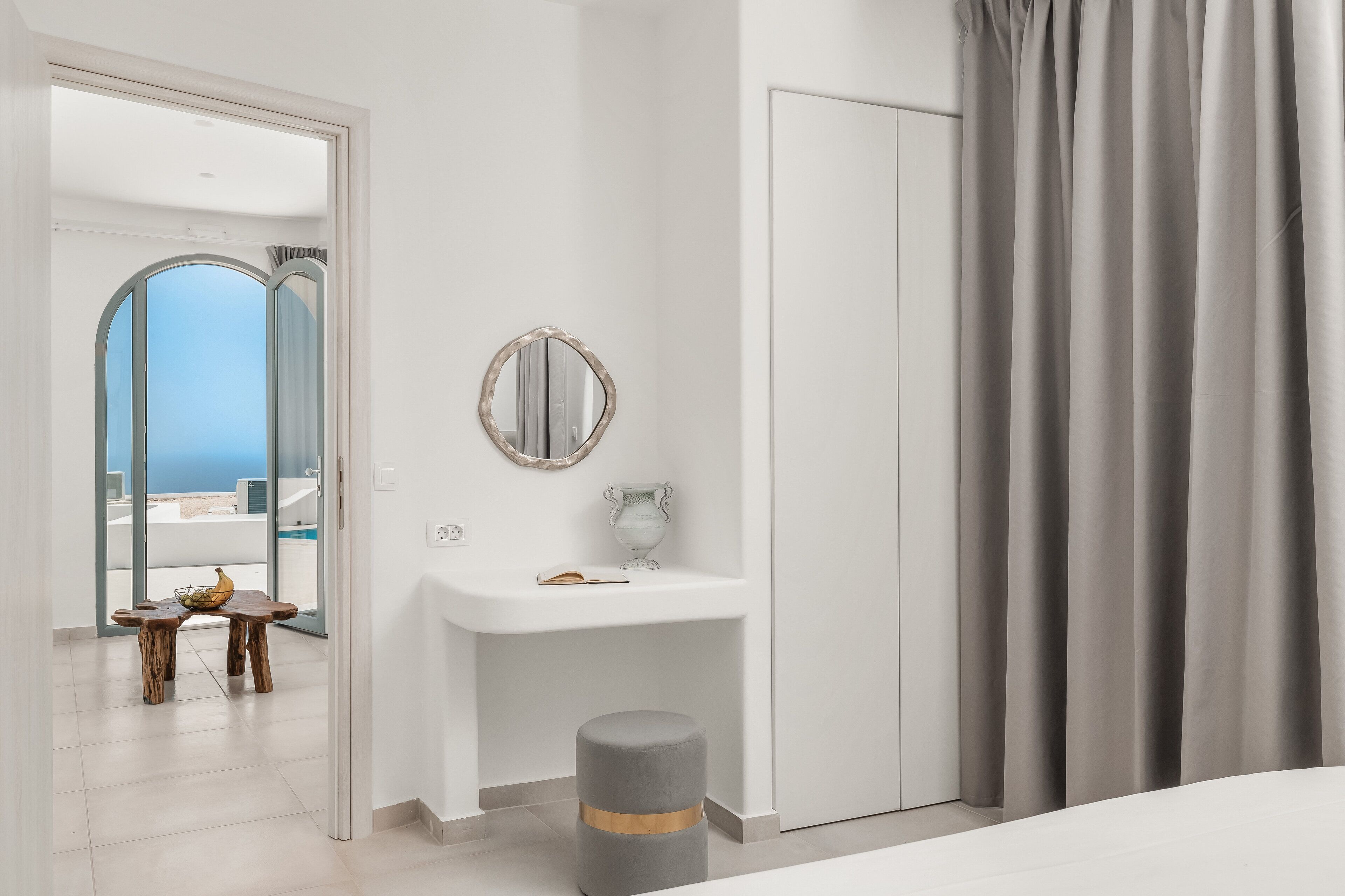 A modern bedroom with a white vanity, a round mirror, and a grey ottoman.