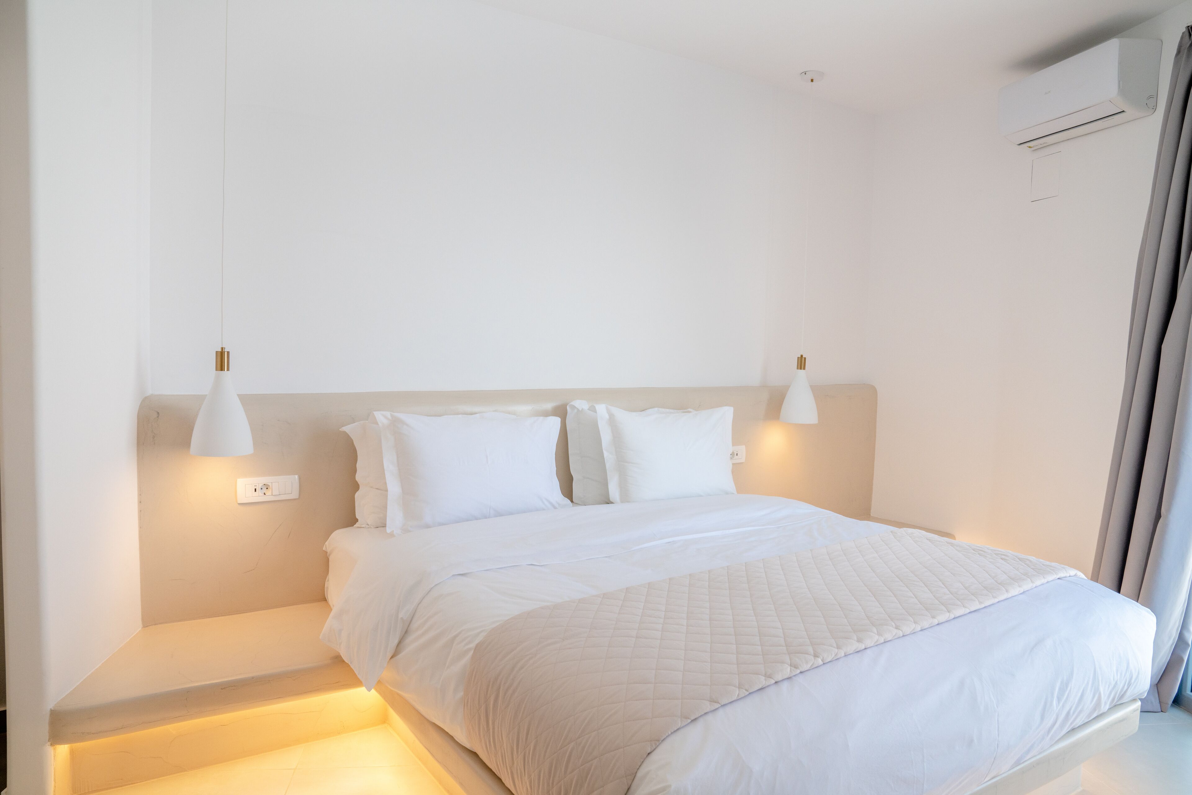 A minimalist bedroom with a large bed, bedside lamps, and an air conditioning unit.